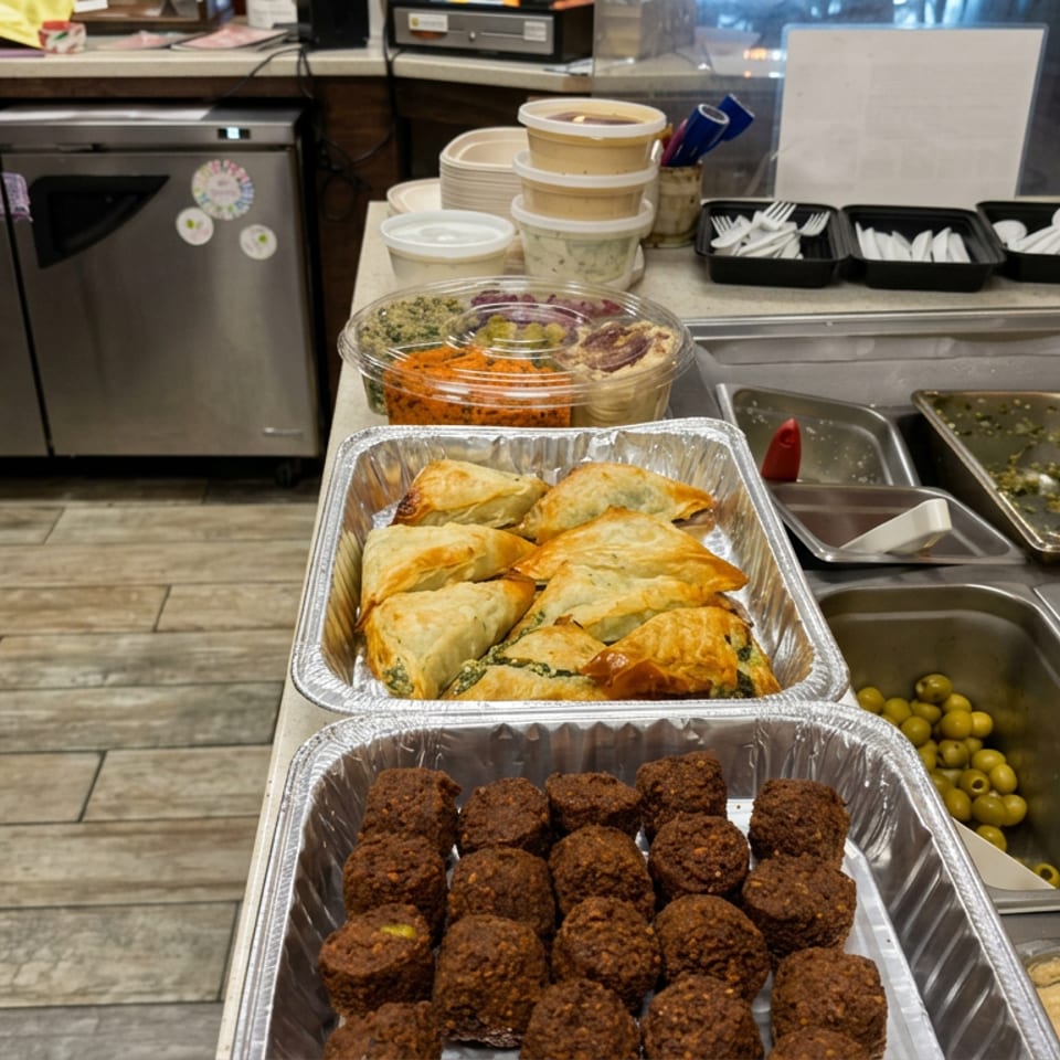 Mediterranean buffet counter displaying falafel, stuffed pastries, hummus, olives, salads, and sauces in serving trays.