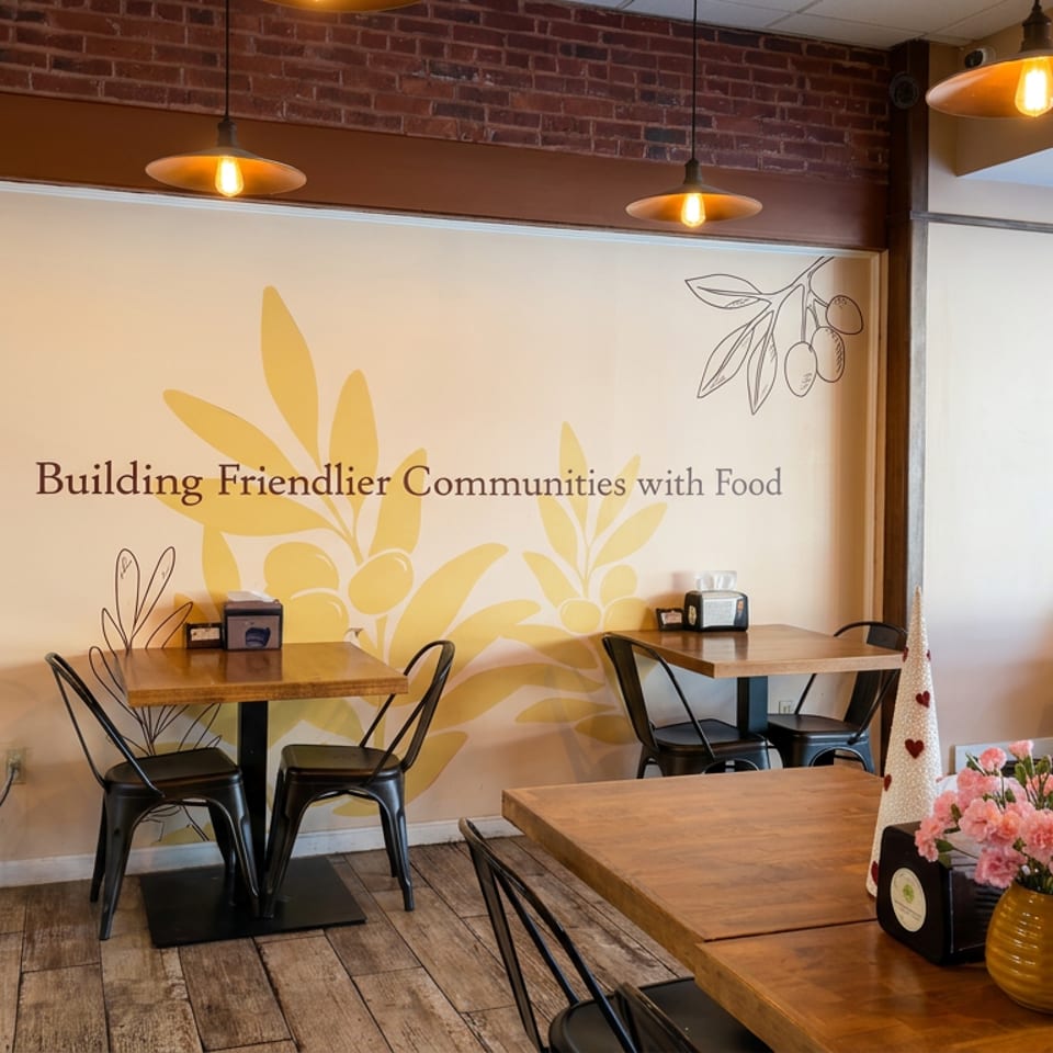Warm restaurant dining area with wooden tables, black chairs, floral centerpiece, pendant lights, and olive branch mural reading “Building Friendlier Communities with Food.”