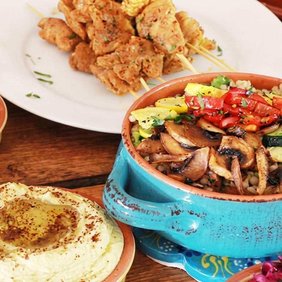 Mediterranean feast featuring hummus, grilled vegetable and grain bowl, fried skewers, dips, bread, and side dishes on a wooden table.