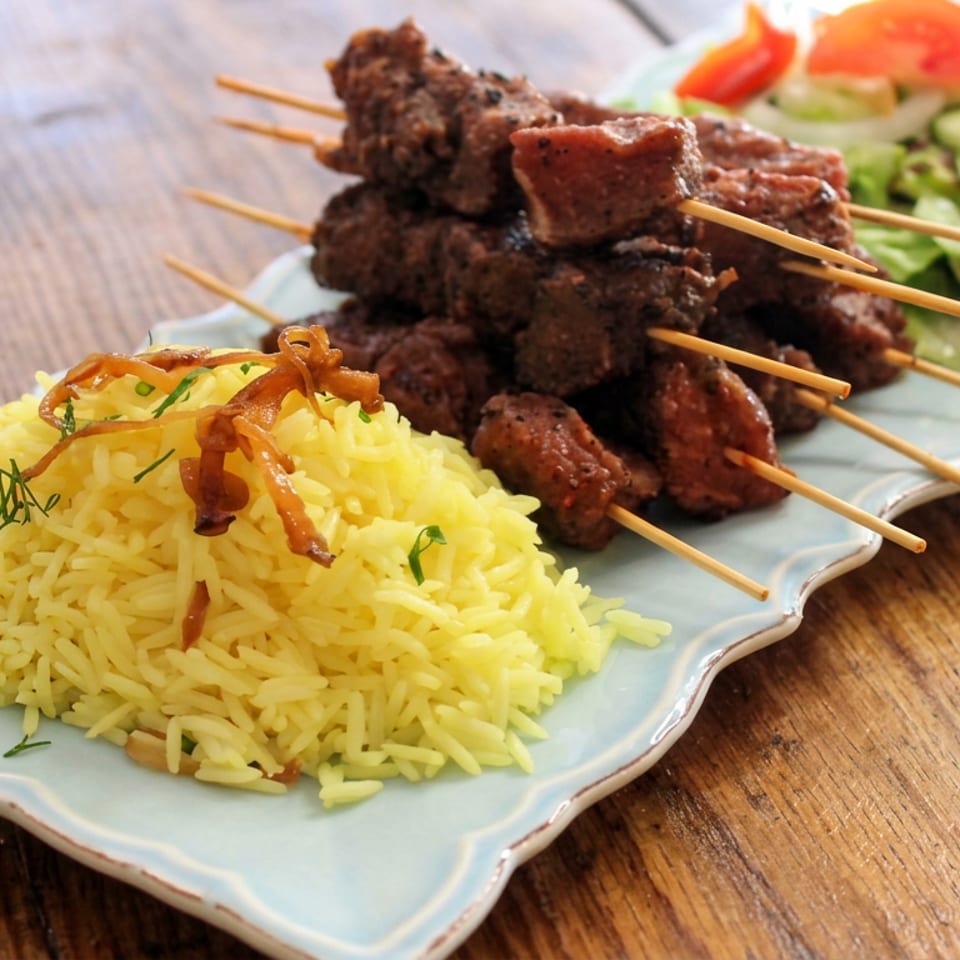 Grilled meat skewers served with yellow rice, fried onions, fresh salad, cucumber, and tomatoes on a decorative plate.