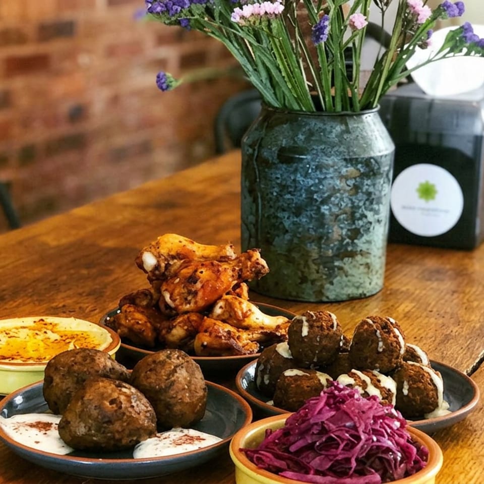 Rustic restaurant table displaying meatballs, chicken wings, creamy dips, purple cabbage slaw, and a vase of flowers.