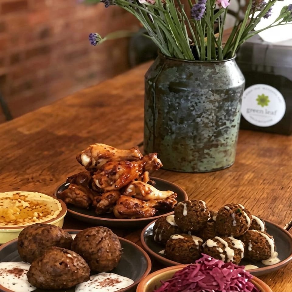 Assorted falafel, glazed chicken wings, hummus, tahini sauce, and shredded purple cabbage arranged on a rustic wooden restaurant table.