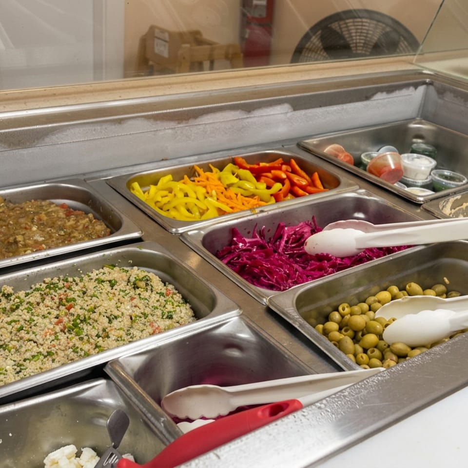 Restaurant salad bar displaying couscous, marinated vegetables, shredded cabbage, olives, hummus, and serving tongs in stainless-steel trays.