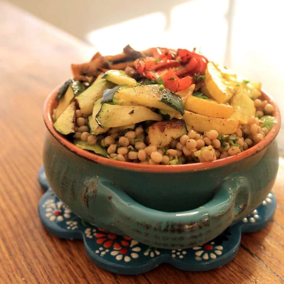 A rustic ceramic bowl holds couscous topped with roasted zucchini, squash, tomatoes, and herbs on a wooden table.