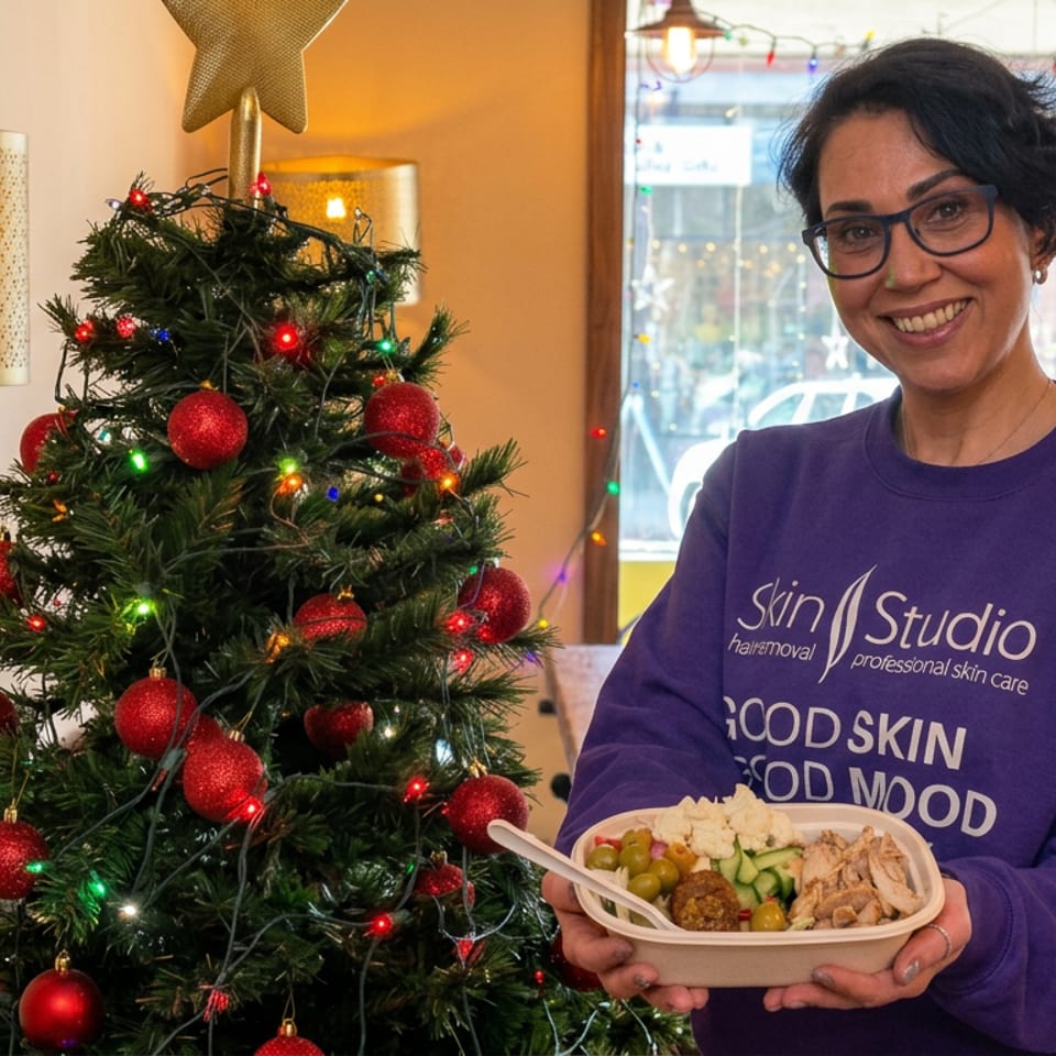 Smiling woman holds a prepared meal beside a decorated Christmas tree inside a warmly lit restaurant.